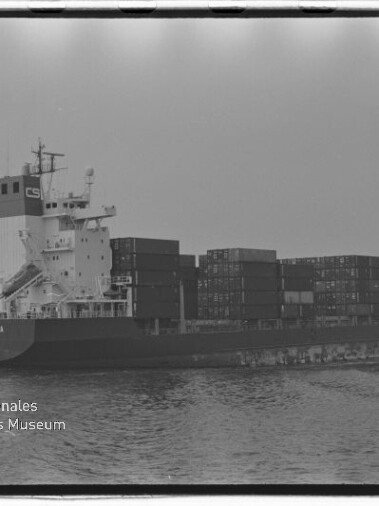 Schwarzweiß-Foto eines beladenen Containerschiffs auf ruhiger See mit mehreren gestapelten Seecontainern, sichtbarem Filmrahmen und Wasserzeichen des Internationalen Maritimen Museums Hamburg unten links.