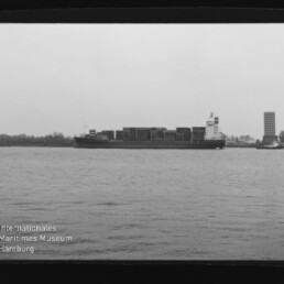 Containerfrachter mit gestapelten Ladecontainern auf ruhigem Wasser, rechts von einem Schlepper begleitet, vor Ufer mit Bäumen und einem hohen Turm; Filmrand mit Aufdruck AGFA APX 25 und Wasserzeichen des Internationalen Maritimen Museums Hamburg sichtbar.