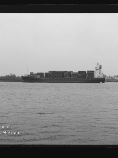 Containerfrachter mit gestapelten Ladecontainern auf ruhigem Wasser, rechts von einem Schlepper begleitet, vor Ufer mit Bäumen und einem hohen Turm; Filmrand mit Aufdruck AGFA APX 25 und Wasserzeichen des Internationalen Maritimen Museums Hamburg sichtbar.
