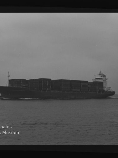 Schwarz-weiß-Foto eines voll beladenen Containerschiffs mit gestapelten Containerreihen, das bei ruhiger See und bedecktem Himmel von links nach rechts fährt; Negativrahmen mit Zahlen und links unten Wasserzeichen „Internationales Maritimes Museum Hamburg“ sichtbar.