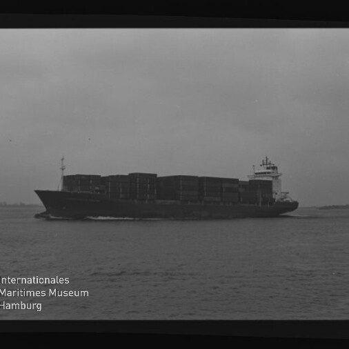 Schwarz-weiß-Foto eines voll beladenen Containerschiffs mit gestapelten Containerreihen, das bei ruhiger See und bedecktem Himmel von links nach rechts fährt; Negativrahmen mit Zahlen und links unten Wasserzeichen „Internationales Maritimes Museum Hamburg“ sichtbar.