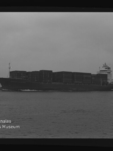 Schwarzweißaufnahme eines großen Containerschiffs, beladen mit gestapelten Containerblöcken, das bei bewölktem Himmel auf ruhiger See von links nach rechts fährt; Filmbord und Wasserzeichen des Internationalen Maritimen Museums Hamburg sind sichtbar.