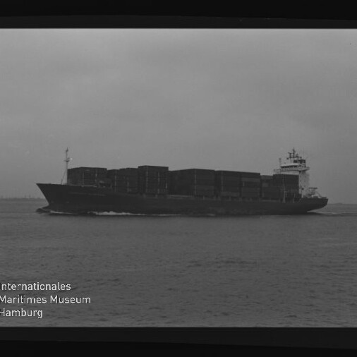 Schwarzweißaufnahme eines großen Containerschiffs, beladen mit gestapelten Containerblöcken, das bei bewölktem Himmel auf ruhiger See von links nach rechts fährt; Filmbord und Wasserzeichen des Internationalen Maritimen Museums Hamburg sind sichtbar.
