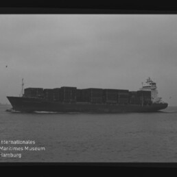 Seitlich abgebildetes Containerschiff mit mehreren übereinander gestapelten Containerreihen, das bei trübem Himmel über das Wasser fährt; das Schwarzweißbild zeigt Filmnegativeinfassung und ein Wasserzeichen des Internationalen Maritimen Museums Hamburg.