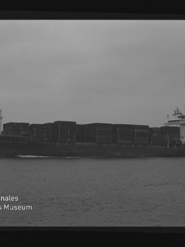 Seitlich abgebildetes Containerschiff mit mehreren übereinander gestapelten Containerreihen, das bei trübem Himmel über das Wasser fährt; das Schwarzweißbild zeigt Filmnegativeinfassung und ein Wasserzeichen des Internationalen Maritimen Museums Hamburg.