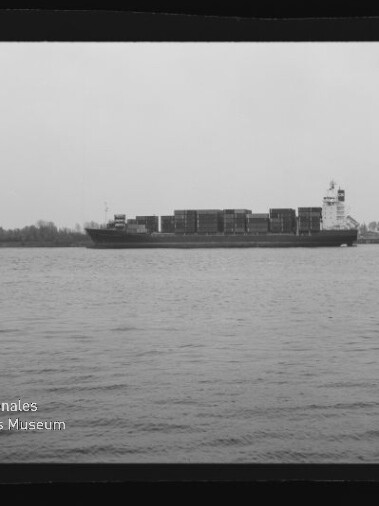 Schwarzweiß-Foto eines großen Containerschiffs mit gestapelten Ladecontainern, das auf ruhigem Wasser vor einer flachen Uferlinie mit Bäumen und einem hohen Lager- oder Bürogebäude fährt; Wasserzeichen des Internationalen Maritimen Museums Hamburg unten links.