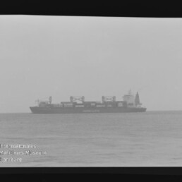 Schwarzweißaufnahme eines Containerschiffs auf ruhiger See am Horizont mit sichtbaren Ladekränen und Schriftzug auf dem Rumpf, Museumswasserzeichen unten links.