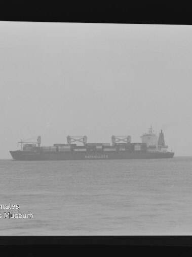 Schwarzweißaufnahme eines Containerschiffs auf ruhiger See am Horizont mit sichtbaren Ladekränen und Schriftzug auf dem Rumpf, Museumswasserzeichen unten links.