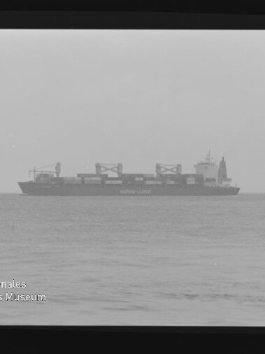 Containerfrachter mit mehreren Deckskränen auf ruhiger See am Horizont unter nebligem Himmel, das Foto im Filmrahmen zeigt unten links das Wasserzeichen des Internationalen Maritimen Museums Hamburg.