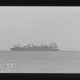Schwarzweißaufnahme eines Containerschiffs auf ruhigem Meer am Horizont, deutlich sichtbar Containerstapel und Ladekräne, mit Filmrand und Wasserzeichen des Internationalen Maritimen Museums Hamburg.