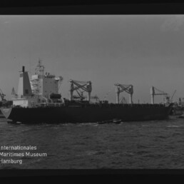 Schwarzweißaufnahme eines großen Frachtschiffs im Hafen mit mehreren markanten Ladekränen, kleinen Begleitbooten auf dem Wasser und einem 'Internationales Maritimes Museum Hamburg'-Wasserzeichen unten links.