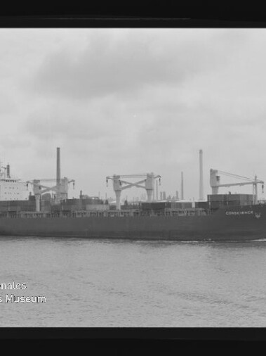 Schwarzweiß-Aufnahme eines Frachtschiffs namens Consequence mit Ladekränen, das auf ruhigem Wasser vor einer industriellen Hafen-Kulisse mit Fabrikschloten fährt.