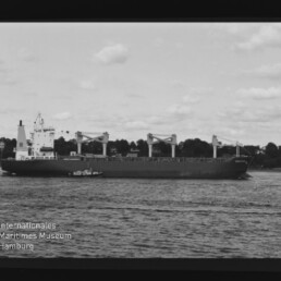 Schwarzweiß-Foto eines großen Frachtschiffs mit vier Ladekränen, begleitet von einem Lotsenboot auf dem Wasser vor einer baumbestandenen Uferlinie; sichtbarer Filmrand und Logo des Internationalen Maritimen Museums Hamburg.