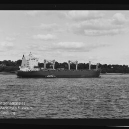 Großes Frachtschiff mit mehreren Ladekränen, begleitet von einem kleinen Schlepper, fährt auf ruhiger See vor einer baumgesäumten Küste unter bewölktem Himmel; im Vordergrund das Logo des Internationalen Maritimen Museums Hamburg.