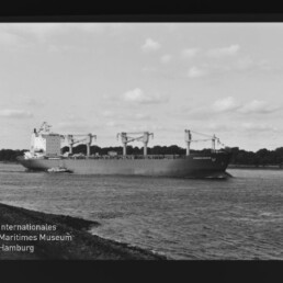 Schwarzweißaufnahme eines großen Frachtschiffs mit mehreren Ladekränen, begleitet von einem Schlepper auf ruhigem Wasser vor einer baumbestandenen Uferlinie und leicht bewölktem Himmel; Filmrand und Museumswasserzeichen sichtbar.