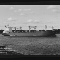 Schwarzweiß-Foto eines großen Frachtschiffs mit drei Deckskränen und einem kleinen Schlepper an seiner Seite, das auf ruhigem Wasser vor einer baumbewachsenen Küstenlinie fährt; Filmrandmarkierungen und Museumswasserzeichen sind im Bild sichtbar.