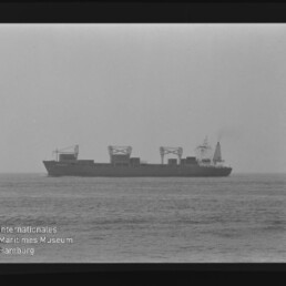 Schwarzweiß-Foto eines Containerschiffs in Seitenansicht auf ruhiger See, sichtbar sind mehrere Ladekräne auf dem Deck, der Filmrand mit Aufdruck 'AGFAPAN 100' und ein Wasserzeichen des Internationalen Maritimen Museums Hamburg im Bild