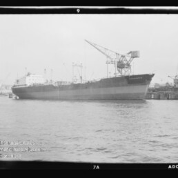 Schwarz-weiß-Fotografie eines großen Frachtschiffs, das an einem Industriehafen mit mehreren hohen Hafenkränen liegt, ruhiges Wasser im Vordergrund und sichtbarer Filmrand sowie Museumswasserzeichen in der Ecke.