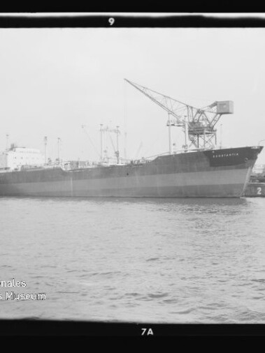 Schwarz-weiß-Fotografie eines großen Frachtschiffs, das an einem Industriehafen mit mehreren hohen Hafenkränen liegt, ruhiges Wasser im Vordergrund und sichtbarer Filmrand sowie Museumswasserzeichen in der Ecke.