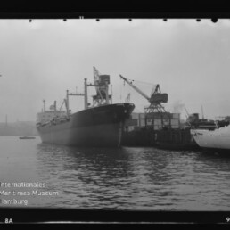 Schwarzweißaufnahme eines großen Frachtschiffs, das am Kai eines Industriehafens liegt, mit Hafenkran und Anlegestelle rechts, ruhigem Wasser im Vordergrund und Wasserzeichen des Internationalen Maritimen Museums Hamburg.