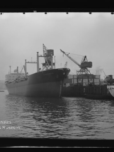 Schwarzweißaufnahme eines großen Frachtschiffs, das am Kai eines Industriehafens liegt, mit Hafenkran und Anlegestelle rechts, ruhigem Wasser im Vordergrund und Wasserzeichen des Internationalen Maritimen Museums Hamburg.
