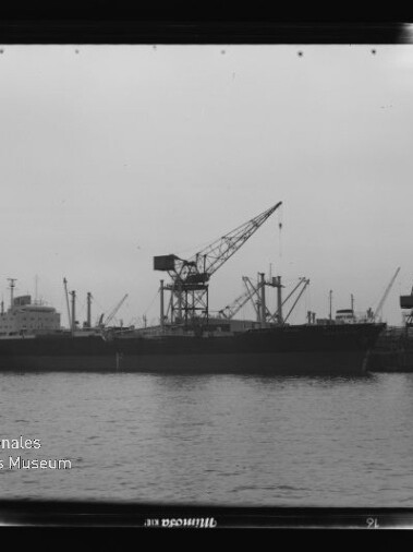 Großes Frachtschiff liegt am Kai eines Industriehafens, umgeben von mehreren hohen Ladekränen, ruhige Wasserfläche im Vordergrund und Wasserzeichen des Internationalen Maritimen Museums Hamburg unten links.
