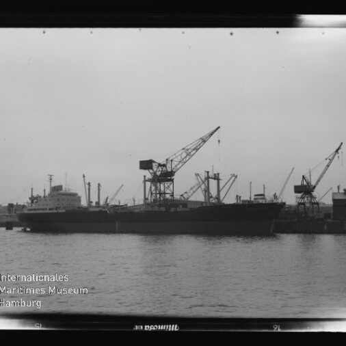 Großes Frachtschiff liegt am Kai eines Industriehafens, umgeben von mehreren hohen Ladekränen, ruhige Wasserfläche im Vordergrund und Wasserzeichen des Internationalen Maritimen Museums Hamburg unten links.