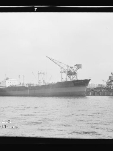 Schwarzweiß-Foto eines großen Frachtschiffs, das am Kai eines Industriehafens mit mehreren Ladekränen und Hafenbauten liegt; im Vordergrund Wasser, sichtbar sind Filmränder und ein Wasserzeichen des Internationalen Maritimen Museums Hamburg.