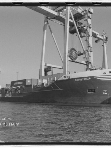 Schwarzweiß-Foto eines Containerschiffs namens Constanza, das längsseits am Kai liegt, mit großen Hafenkränen über dem Deck, sichtbaren Containerstapeln und Wasser im Vordergrund; unten links das Logo des Internationalen Maritimen Museums Hamburg.