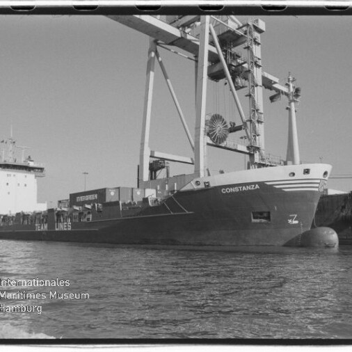 Schwarzweiß-Foto eines Containerschiffs namens Constanza, das längsseits am Kai liegt, mit großen Hafenkränen über dem Deck, sichtbaren Containerstapeln und Wasser im Vordergrund; unten links das Logo des Internationalen Maritimen Museums Hamburg.