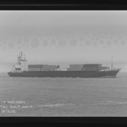 Schwarzweiß-Profilaufnahme eines Containerschiffs auf ruhiger See mit gestapelten Ladecontainern, gerahmter Filmnegativscan mit sichtbarem Museumswasserzeichen links unten.