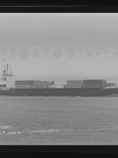 Schwarzweiß-Profilaufnahme eines Containerschiffs auf ruhiger See mit gestapelten Ladecontainern, gerahmter Filmnegativscan mit sichtbarem Museumswasserzeichen links unten.