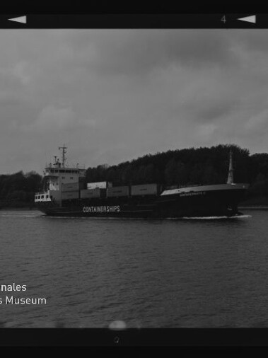 Schwarzweißaufnahme eines Containerschiffs mit der Aufschrift CONTAINERSHIPS, das auf einem ruhigen Fluss oder Kanal entlangfährt, vor bewaldetem Ufer und bewölktem Himmel; Museumslogo unten links.