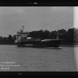 Schwarzweißfoto eines Containerschiffs mit der Aufschrift CONTAINERSHIPS, das mehrere Container geladen vor einem baumbestandenen Ufer auf dem Wasser fährt.