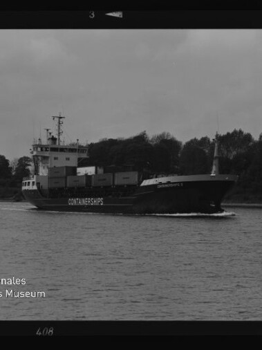 Schwarzweißfoto eines Containerschiffs mit der Aufschrift CONTAINERSHIPS, das mehrere Container geladen vor einem baumbestandenen Ufer auf dem Wasser fährt.
