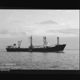 Schwarz-weiß-Foto eines Frachtschiffs mit mehreren Ladekränen und markanten Aufbauten, das ruhig auf dem Meer fährt; links unten sichtbar das Logo und der Schriftzug des Internationalen Maritimen Museums Hamburg.