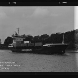 Schwarzweißaufnahme eines Containerschiffs mit aufgestapelten Containerboxen und großem Schriftzug 