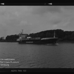Schwarzweiß-Filmaufnahme eines Containerschiffs mit aufgestapelten Containern, das auf einem ruhigen Fluss vor einem bewaldeten Ufer unter wolkigem Himmel fährt; Filmrandbeschriftungen 