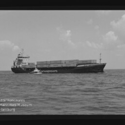 Schwarzweiß-Foto eines Containerschiffs mit aufgestapelten Containerboxen, begleitet von einem kleineren Lotsenboot auf offener See; sichtbarer Filmrand mit Beschriftung ‚AGFA PAN 100‘ und Wasserzeichen des Internationalen Maritimen Museums Hamburg.