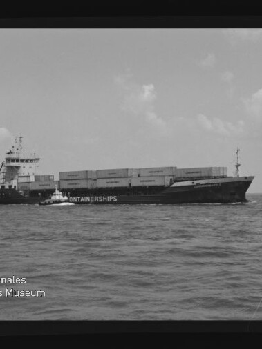 Schwarzweiß-Foto eines Containerschiffs mit aufgestapelten Containerboxen, begleitet von einem kleineren Lotsenboot auf offener See; sichtbarer Filmrand mit Beschriftung ‚AGFA PAN 100‘ und Wasserzeichen des Internationalen Maritimen Museums Hamburg.