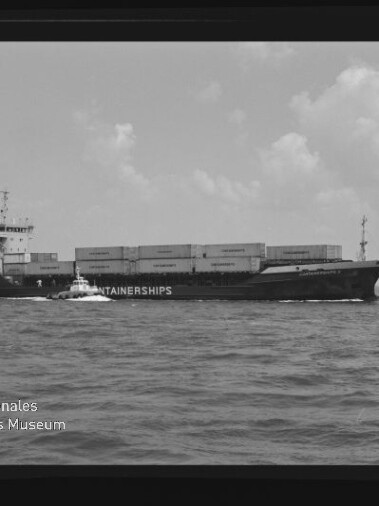 Schwarzweiß-Foto eines beladenen Containerschiffs mit Aufschrift CONTAINERSHIPS, neben dem ein kleines Lotsenboot fährt; ruhige See, vereinzelte Wolken am Horizont sowie sichtbarer Filmrand und Wasserzeichen des Internationalen Maritimen Museums Hamburg