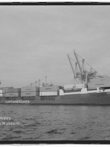 Schwarzweißbild eines beladenen Containerschiffs mit dem Schriftzug „CONTAINERSHIPS“, das im Hafen neben großen Hafenkranen liegt; unten links das Logo des Internationalen Maritimen Museums Hamburg.