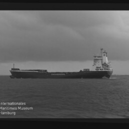 Schwarzweiß-Aufnahme eines Containerschiffs mit der Aufschrift „CONTAINERSHIPS“, das bei bewölktem Himmel von links nach rechts auf dem ruhigen Meer fährt; links unten im Bild das Logo und der Schriftzug des Internationalen Maritimen Museums Hamburg sichtbar.