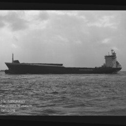 Schwarzweiß-Foto eines seitlich gezeigten Containerschiffs auf ruhiger See unter bewölktem Himmel, mit sichtbarem Filmrand und Schriftzug „AGFA APX 100“ sowie Wasserzeichen des Internationalen Maritimen Museums Hamburg.