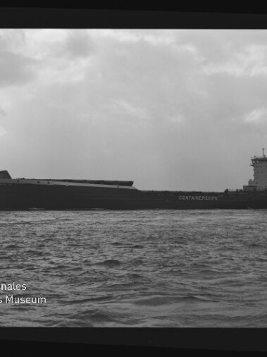 Schwarzweiß-Foto eines seitlich gezeigten Containerschiffs auf ruhiger See unter bewölktem Himmel, mit sichtbarem Filmrand und Schriftzug „AGFA APX 100“ sowie Wasserzeichen des Internationalen Maritimen Museums Hamburg.