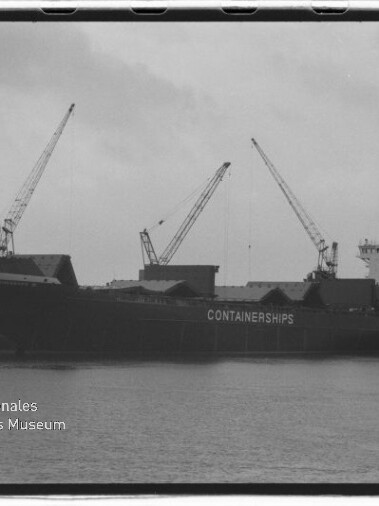 Schwarzweißaufnahme eines großen Containerschiffs mit der Aufschrift „CONTAINERSHIPS“, festgemacht am Kai neben mehreren Hafenkränen, Wasserfläche im Vordergrund und Museumslogo unten links.