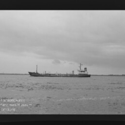 Schwarzweißfoto eines Frachtschiffs auf ruhiger See vor einer flachen Küstenlinie unter bewölktem Himmel; Filmrahmen und das Wasserzeichen „Internationales Maritimes Museum Hamburg“ sind unten links sichtbar.