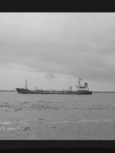 Schwarzweißfoto eines Frachtschiffs auf ruhiger See vor einer flachen Küstenlinie unter bewölktem Himmel; Filmrahmen und das Wasserzeichen „Internationales Maritimes Museum Hamburg“ sind unten links sichtbar.