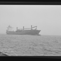 Schwarzweißaufnahme eines Frachtschiffs mit drei Ladekränen auf offenem, leicht nebligem Meer; bewegte See im Vordergrund und unten links das Wasserzeichen des Internationalen Maritimen Museums Hamburg.
