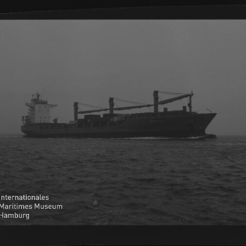 Schwarzweißaufnahme eines Frachtschiffs mit drei Ladekränen und achterlichem Aufbauteil, das bei ruhiger See unter bedecktem Himmel auf dem Meer fährt; unten links sichtbares Logo des Internationalen Maritimen Museums Hamburg.